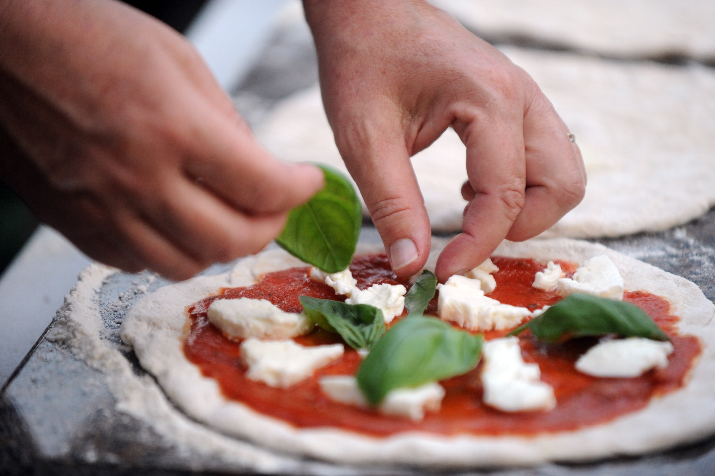 Hands laying fresh basil and mozzarella onto a hand-stretched pizza